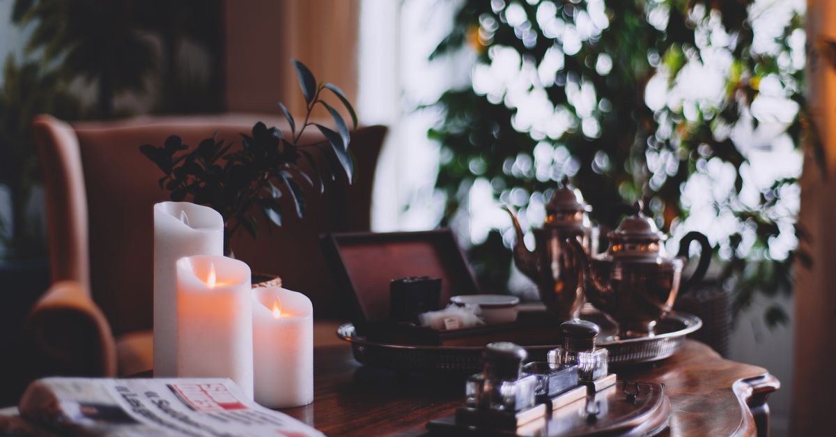Flaming wax candles near metal teapots on tray and cozy armchair in house room
