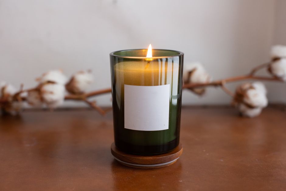 A lit candle in a green glass jar with a blank label, set against a cozy cotton branch backdrop.