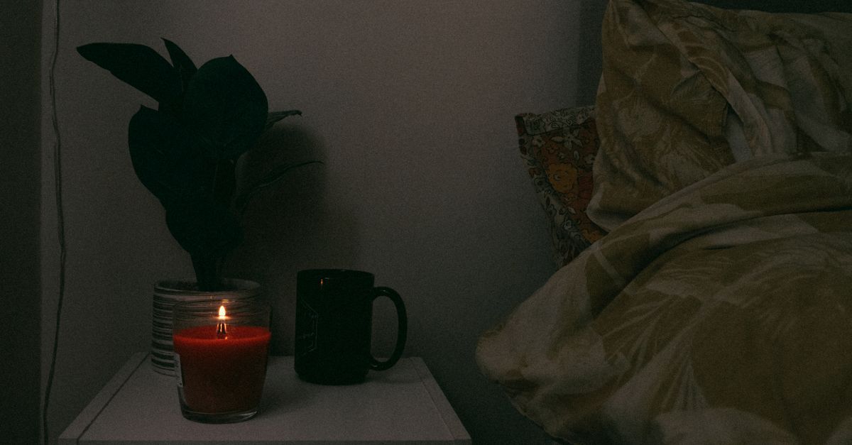 A cozy nightstand scene with a candle, mug, and plant beside a bed, creating a warm atmosphere.
