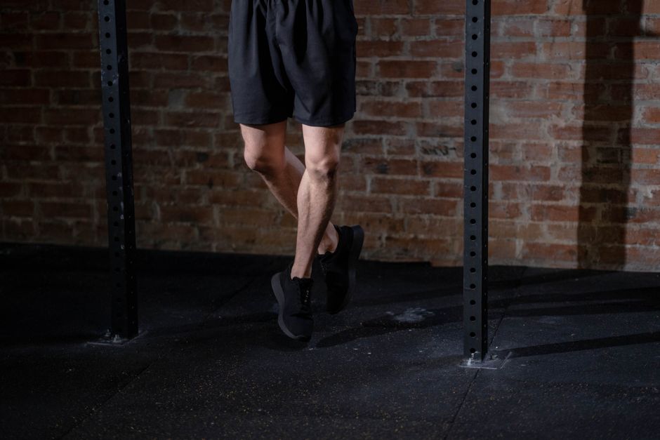A person in black shorts and sneakers stands on a gym floor with brick wall background.