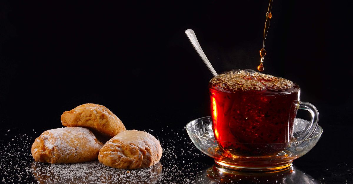 Warm tea being poured into a glass cup with powdered sugar biscuits on the side.