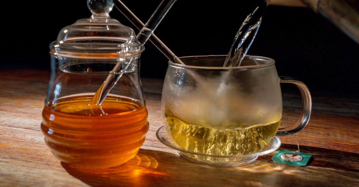 A steaming cup of tea being poured next to a jar of honey, set on a warm wooden table.