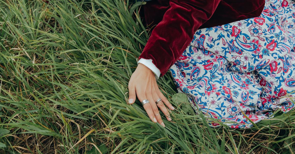 A woman's hand with silver rings rests gently on green grass, wearing a floral patterned skirt.