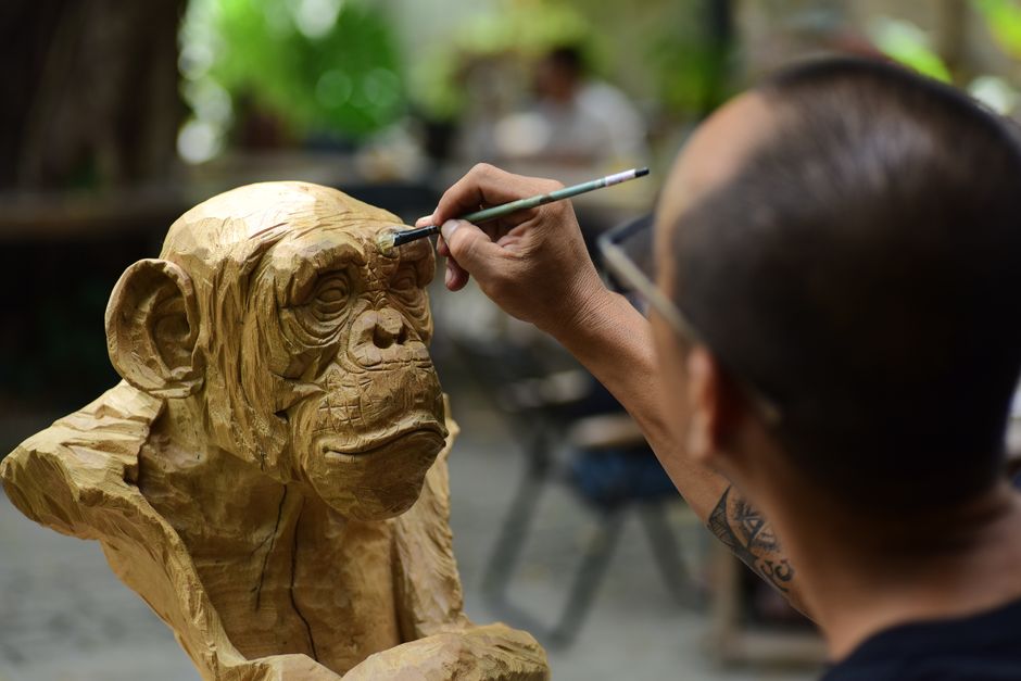 Artisan carving intricate details on a wooden chimpanzee sculpture outdoors.